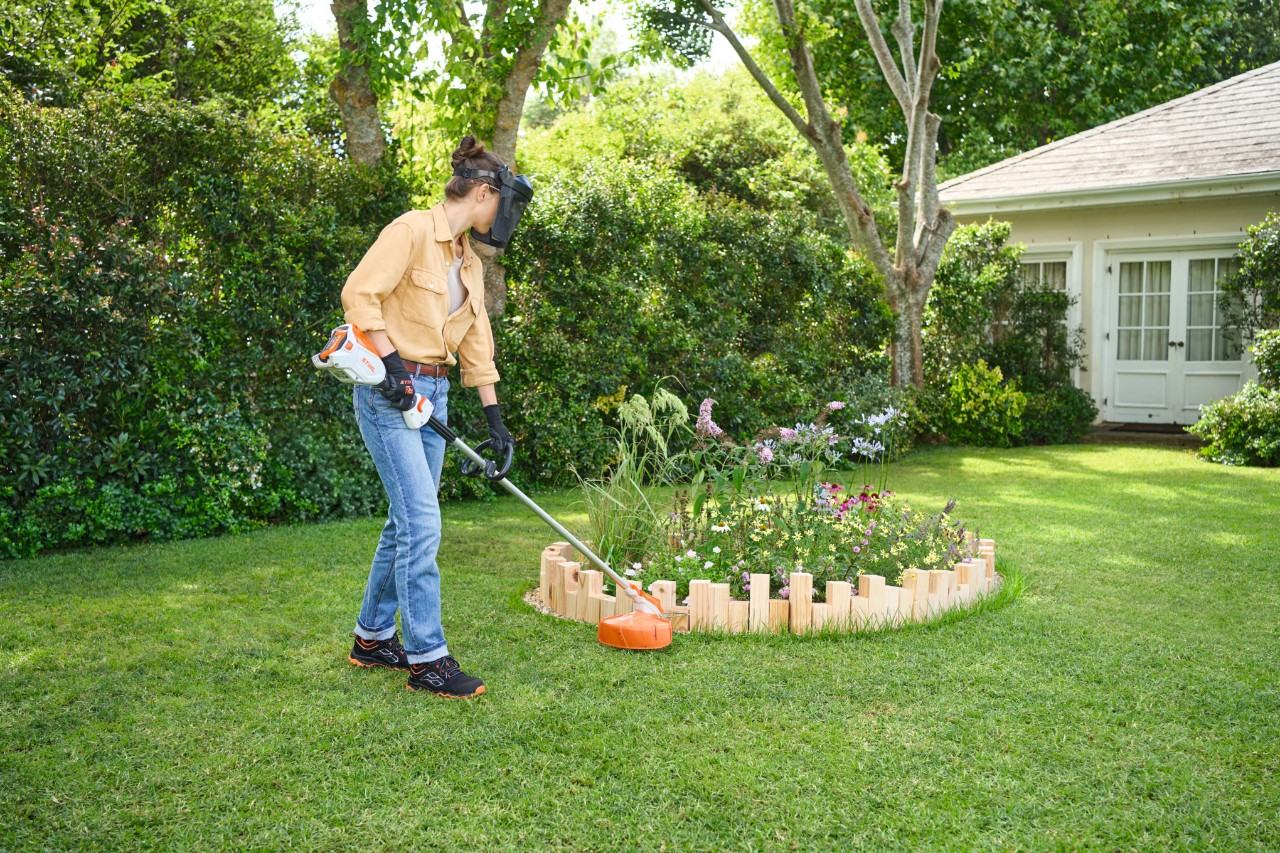 Eine Frau schneidet mit dem STIHL Freischneider FSA 60 R das hochgewachsene Gras um einer Beeteinfassung.