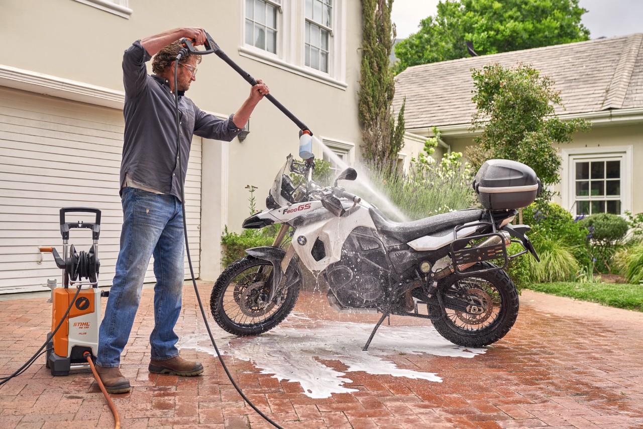 Un hombre utiliza la hidrolimpiadora STIHL RE 130 plus para lavar la moto con agua a presión y detergente de vehículos. 