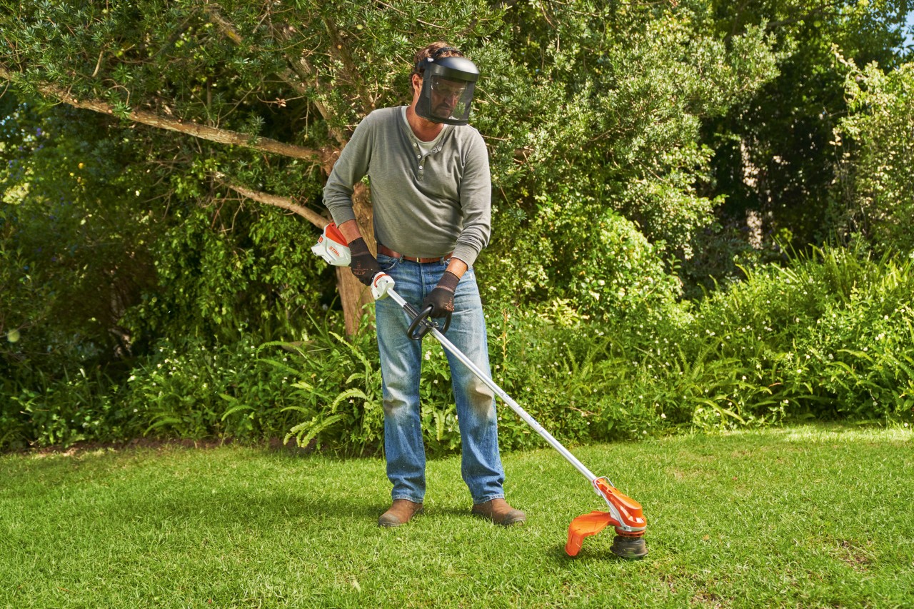 Un hombre con protector facial corta el césped en un jardín con la desbrozadora de batería STIHL FSA 60