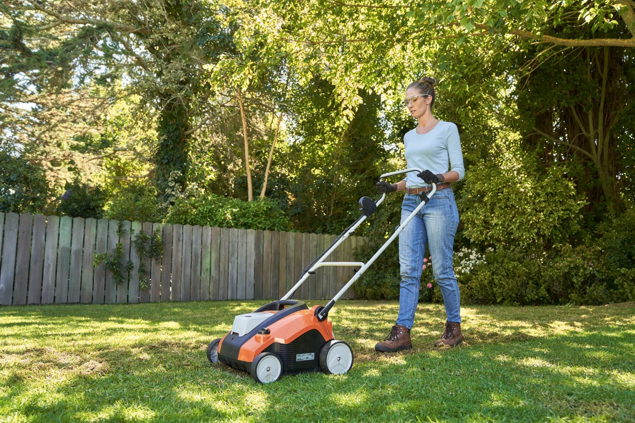 Una mujer airea el césped con el escarificador STIHL RLA 240 en el jardín, equipada con gafas y guantes de trabajo.