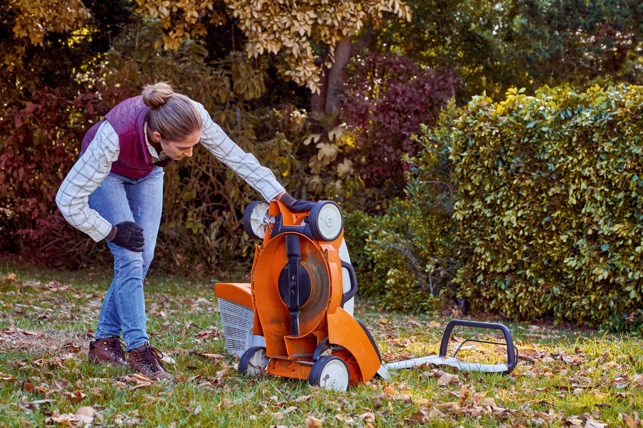 Una mujer revisa la parte inferior de un cortacésped STIHL RMA 339 C en un jardín durante el cuidado del césped en otoño