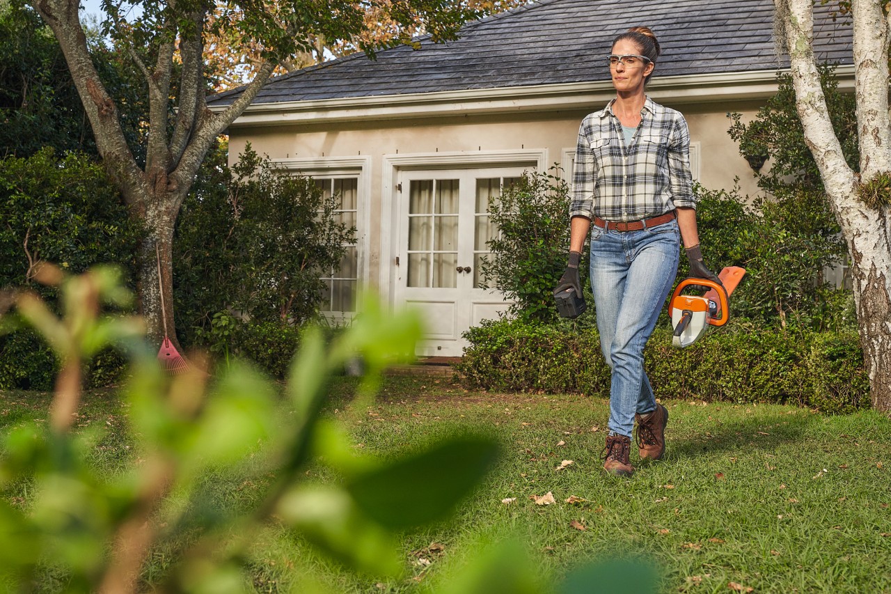  Una mujer con gafas de protección y guantes camina por un jardín con un cortasetos STIHL en la mano izquierda