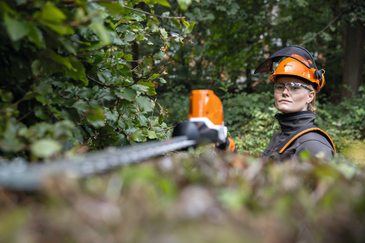 Mujer rubia con equipo de protección cortando un seto de haya