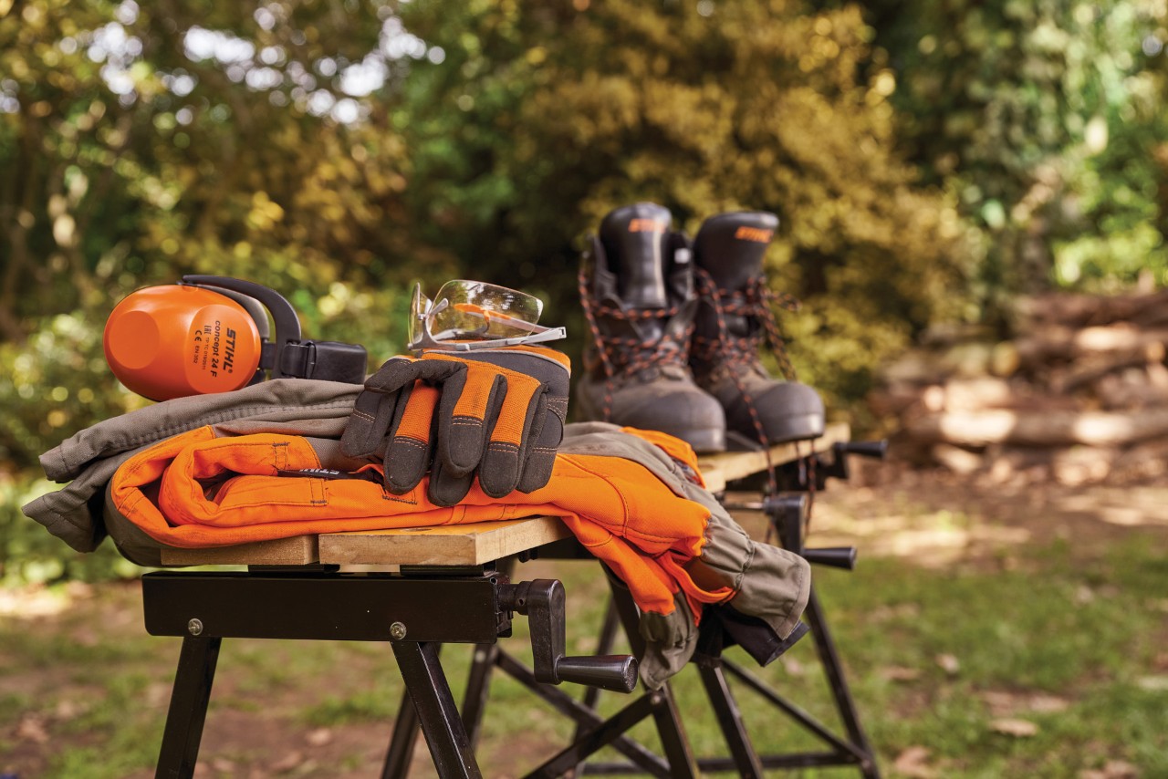 Protección auditiva, botas, gafas de seguridad y guantes de protección STIHL en un banco de trabajo de un jardín.