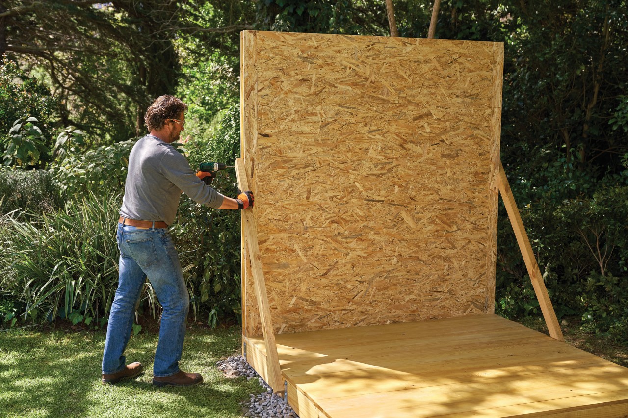 Cómo hacer una caseta de jardín: un hombre monta una pared premontada en una base utilizando soportes temporales.