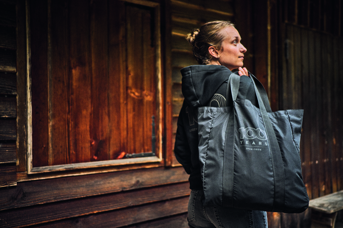 Mujer con la sudadera "STIHL 100" de la colección aniversario frente a una cabaña de madera.
