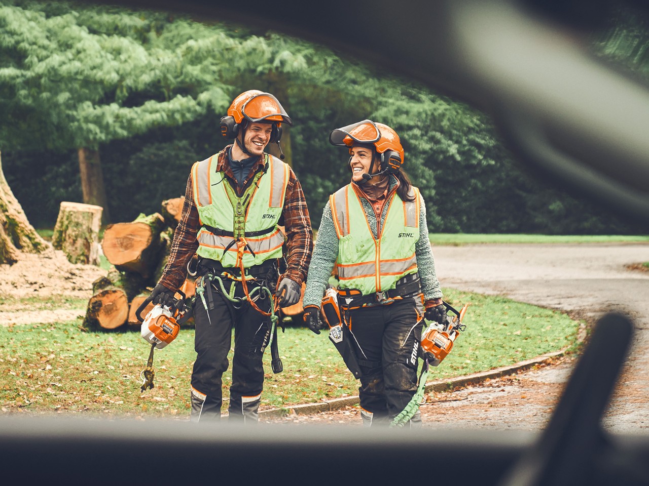 Un hombre y una mujer con motosierras sin cable STIHL y una podadora sin cable STIHL caminan sonrientes hacia un coche.