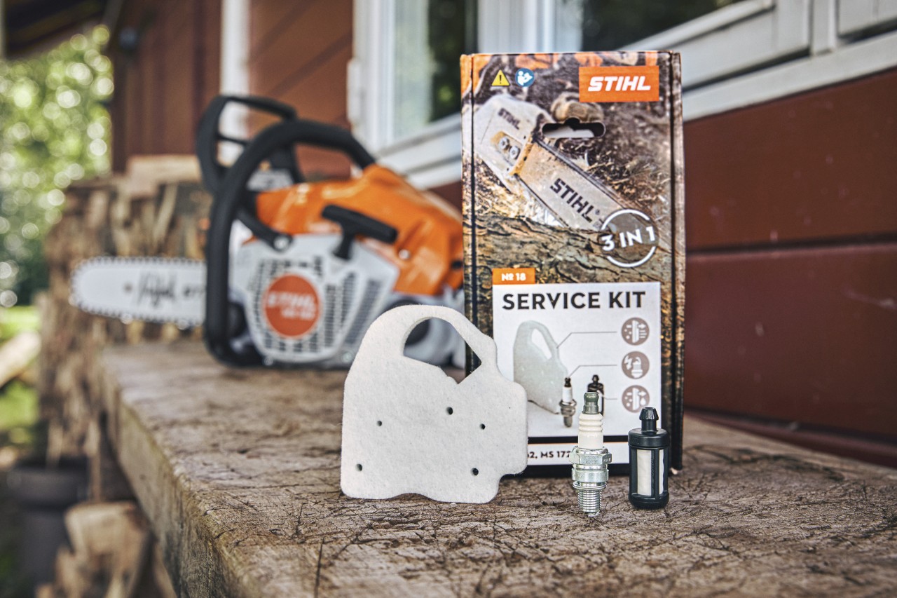 Kit de servicio STIHL 18 en un banco de trabajo, delante los componentes individuales filtro de aire de fieltro, bujía y filtro de combustible