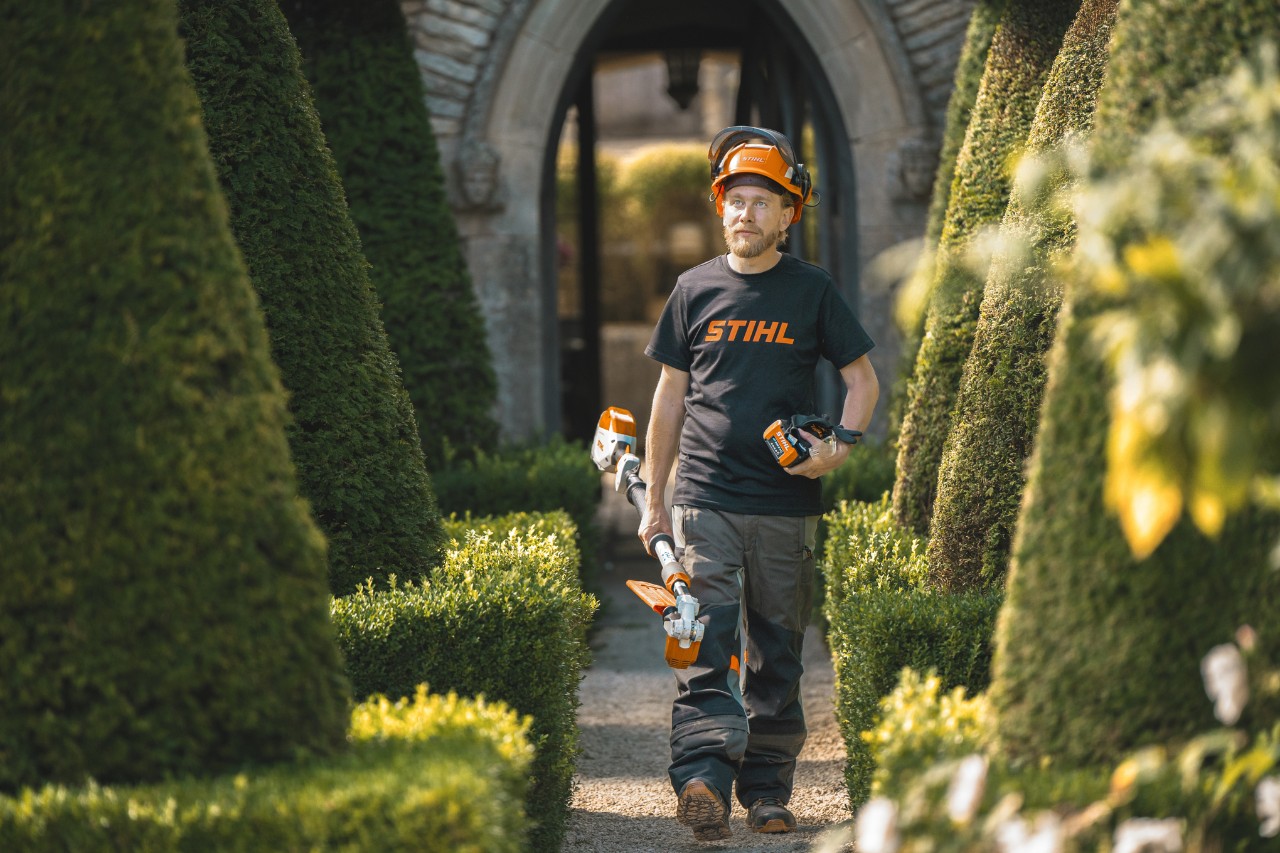 El jardinero jefe Andrew Wain recorre un camino del jardín con un casco de seguridad, una camiseta STIHL y una máquina de batería STIHL con batería independiente en la mano
