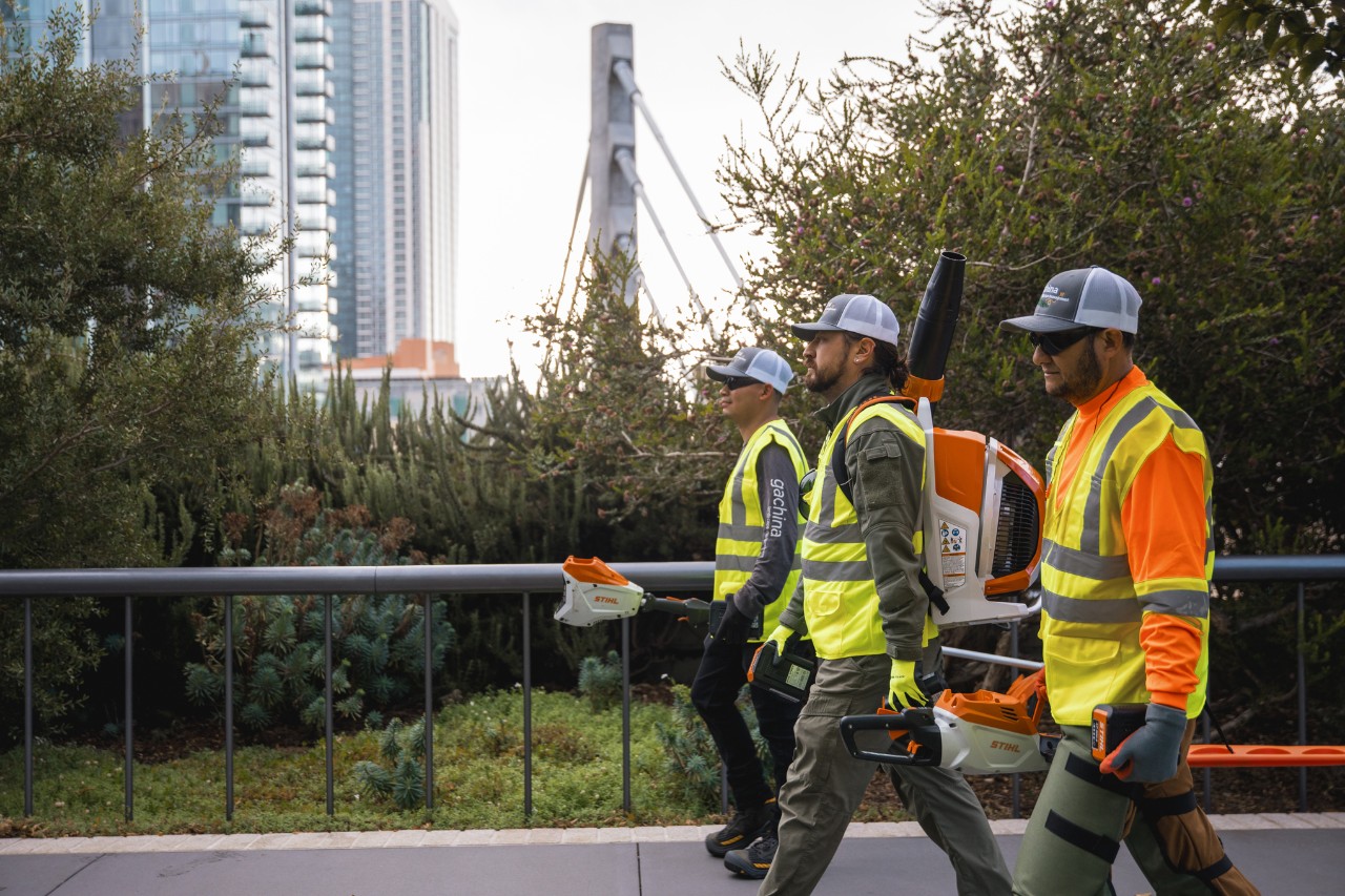Tres jardineros recorren una ciudad con ropa de trabajo y máquinas STIHL.