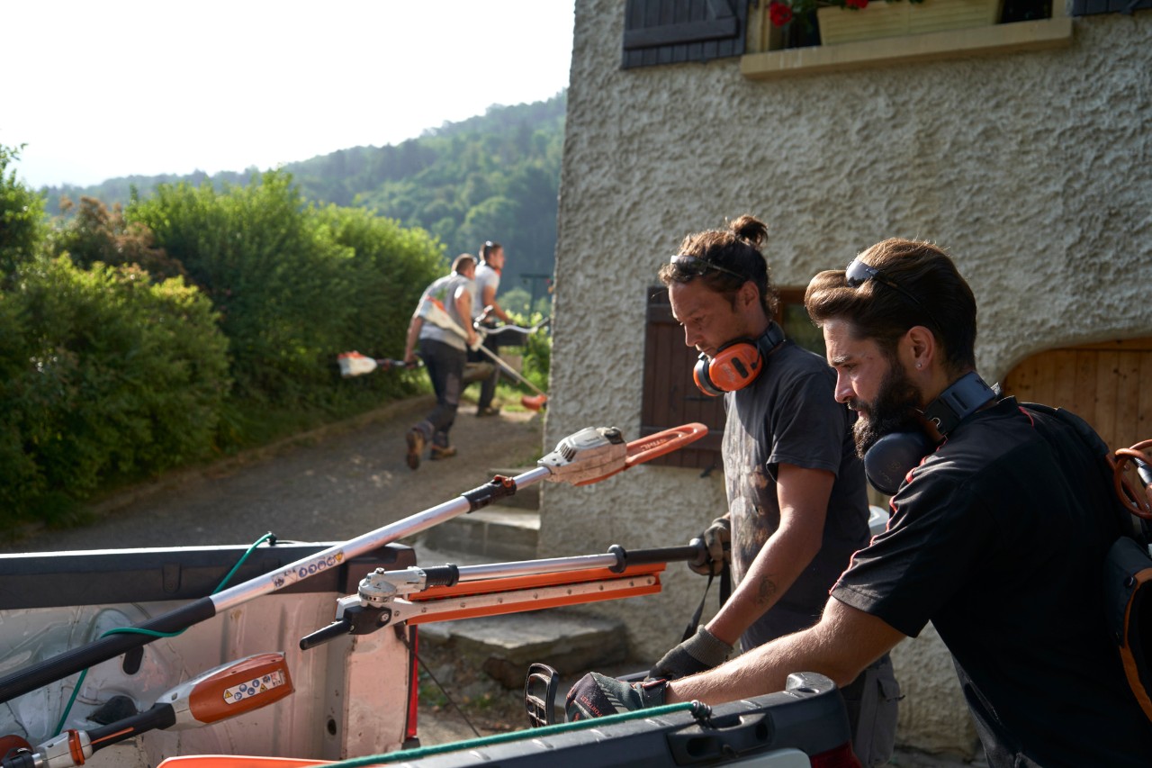Dos jardineros paisajistas descargan las máquinas de batería STIHL de un coche; en segundo plano hay otros trabajadores 