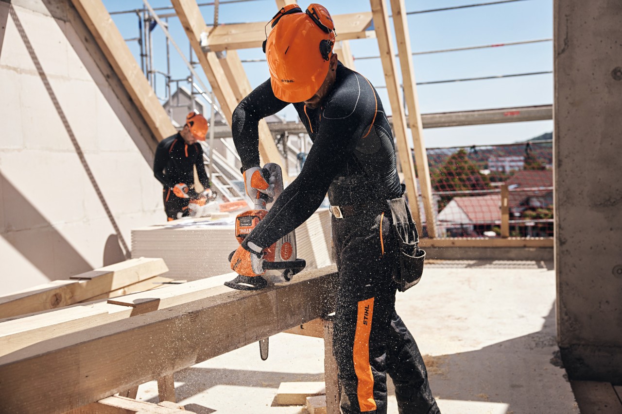 Un profesional de STIHL en una estructura del techo sierra una viga de madera con una motosierra de batería, al fondo otro trabajador maneja una motosierra de batería