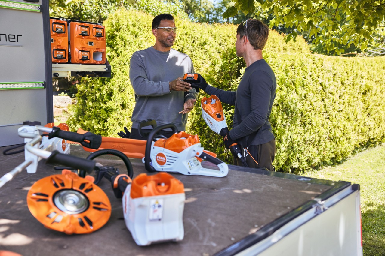 Un hombre entrega una batería STIHL a su compañero, ambos se encuentran detrás de la superficie de carga de un vehículo, entre ellos el bottTainer y una motosierra de batería STIHL, así como otras máquinas