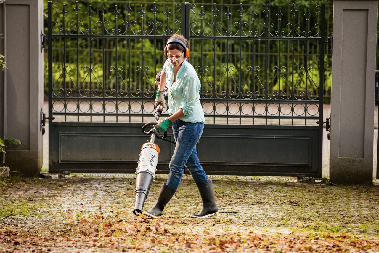 Una mujer con gafas de seguridad y protección auditiva utiliza un motor Combi STIHL KM 131 R con accesorio soplador para recoger las hojas